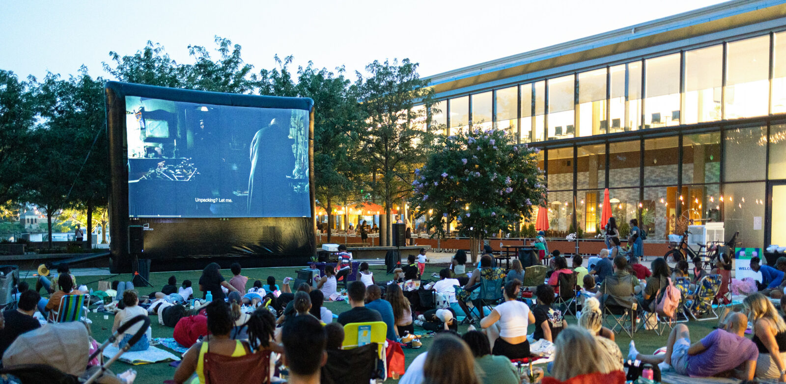 A group of people sits on a lawn and views an outdoor movie on a big screen.