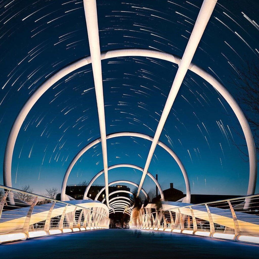 Looking up from a bridge in Yards Park, a time-lapse view of the night sky shows stars blurring in a circular motion.